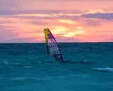 Ein Windsurfer gleitet bei Sonnenuntergang über das Meer. Die bunten Wolken spiegeln sich im Wasser. Ein perfekter Tag für Windsurfen Mallorca.