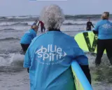 Ältere Dame mit grauem Haar und Surfbrett, bereit für Hvide Sande Surfen. Mehrere Personen in blauen Shirts mit "Blue Spirit" Aufdruck im Wasser.