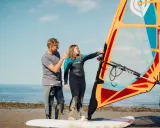Vater erklärt Tochter das Windsurfen NRW. Sie stehen am Strand mit einem Segel und Board.