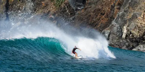 Surfer reitet eine grüne Welle vor einer felsigen Küste. Perfekt für die Surf-App, um die nächste Session zu planen.