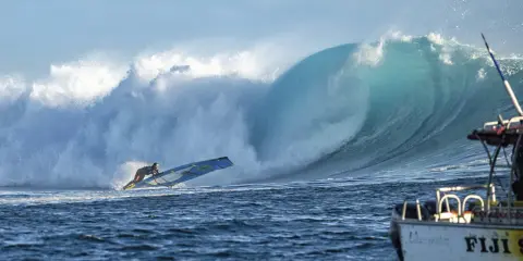 Sarah Hauser surft auf einer riesigen Welle, die sich hinter ihr bricht. Ein Boot mit der Aufschrift "Fiji S" liegt im Vordergrund.