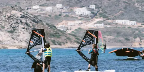 Kinder lernen naxos surfen. Zwei junge Windsurfer üben auf dem Meer, im Hintergrund weiße Häuser am Hang.