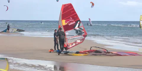 Windsurfer bereiten sich am Strand von Pozo Izquierdo, Gran Canaria, auf das Wasser vor. Mehrere bunte Segel und Boards liegen am Sand.