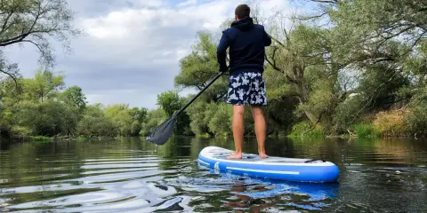 Mann auf SUP-Board auf ruhigem Fluss, umgeben von grünen Bäumen. Perfekt, um zu sehen, wo man stand up paddeln kann.