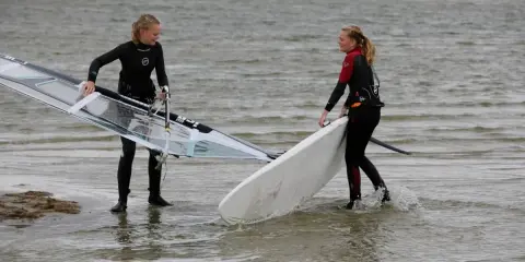 Zwei Mädchen bereiten sich auf das Windsurfen am IJsselmeer vor, tragen Neoprenanzüge und halten ihre Ausrüstung.