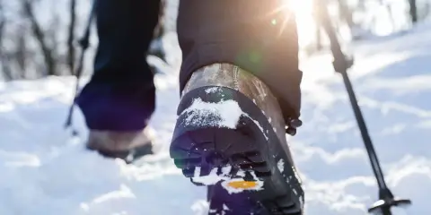 Schneeschuhwandern Schuhe mit Schnee bedeckt, während die Sonne durch die Bäume scheint. Ein Wanderstock ist im Hintergrund zu sehen.