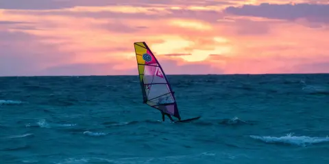 Ein Windsurfer gleitet bei Sonnenuntergang über das Meer. Die bunten Wolken spiegeln sich im Wasser. Ein perfekter Tag für Windsurfen Mallorca.