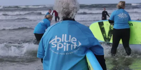 Ältere Dame mit grauem Haar und Surfbrett, bereit für Hvide Sande Surfen. Mehrere Personen in blauen Shirts mit "Blue Spirit" Aufdruck im Wasser.
