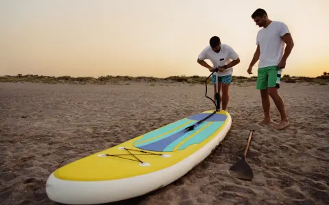Zwei Männer bereiten ein SUP-Board am Strand vor. Einer pumpt es auf, fragt sich: "Sup, wieviel Bar?