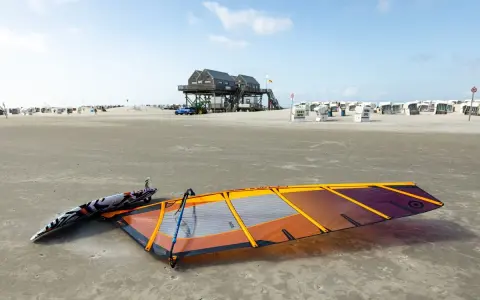 Windsurfen St. Peter-Ording: Segel und Brett liegen am Strand, bereit für den nächsten Ritt auf den Wellen. Strandkörbe und das markante Stelzenhaus im Hintergrund.