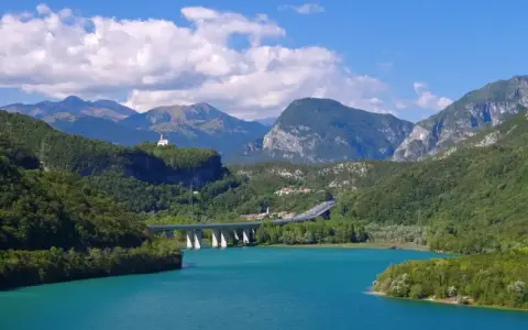 Türkises Wasser des Lago di Cavazzo umgeben von grünen Bergen. Eine Brücke überspannt den See, im Hintergrund eine kleine Kirche auf einem Hügel.