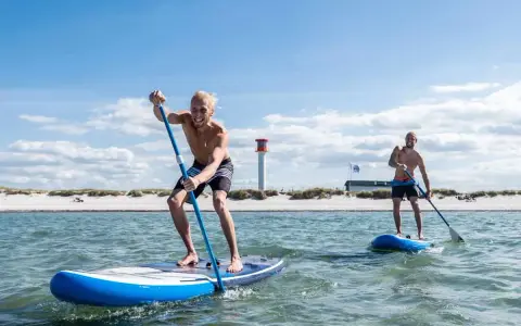 Zwei Männer genießen SUP auf der Nordsee. Sonnenschein, Strand und ein Leuchtturm im Hintergrund.