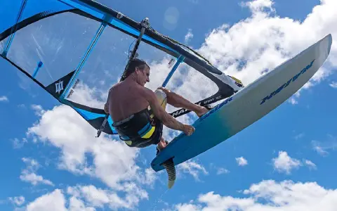 Ein Windsurfer mit einem Freeride Windsurfboard springt hoch in den blauen Himmel mit Wolken.