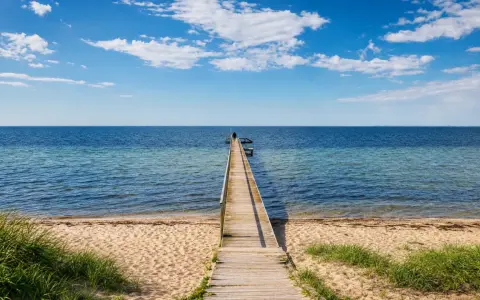 Holzsteg führt ins Meer am Ringkøbing Fjord. Eine Person steht am Ende des Stegs. Blauer Himmel mit Wolken.