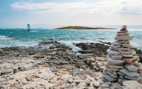 Ein Windsurfer gleitet über das türkisfarbene Meer vor Kroatien. Steinformationen säumen die felsige Küste.