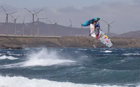 Windsurfer Britt Dunkerbeck springt hoch über den Wellen, im Hintergrund Windräder.