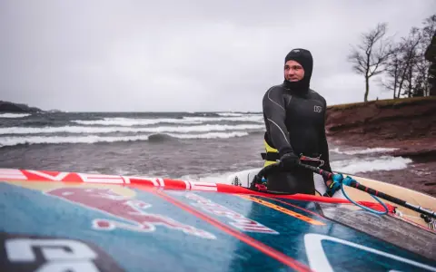 Mann in Neoprenanzug mit Windsurfbrett am Strand. Kaltes Wetter für Windsurfen in Deutschland.