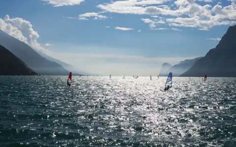 Windsurfer tummeln sich auf dem glitzernden Gardasee vor einer Bergkulisse.