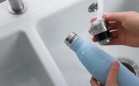 Hände reinigen eine hellblaue Trinkflasche unter fließendem Wasser im Waschbecken.