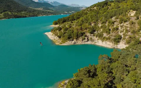 Türkisfarbener Lac de Monteynard mit einem Windsurfer, umgeben von bewaldeten Hügeln und Bergen.