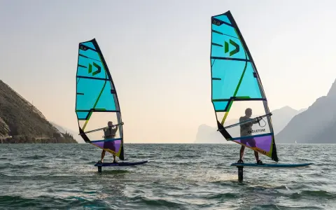 Zwei Personen beim Windsurfen in Italien. Die türkisfarbenen Segel sind im Gegenlicht zu sehen, im Hintergrund Berge.