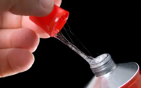 Ein Finger hält die rote Kappe eines Tubenklebers, aus dem zäher, glitzernder Kleber in Fäden heraustropft.