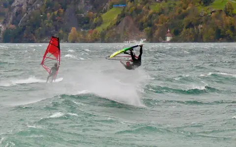 Windsurfer vollführt Sprung am Starnberger See. Ein weiterer Surfer mit rotem Segel im Hintergrund.