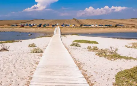 Holzsteg führt über Sand zu einem ruhigen Gewässer. Dahinter parken Autos, die auf einen Ausflug am **mari ermi** warten.