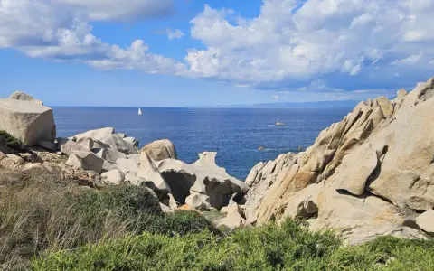 Felsformationen am Capo Testa mit Blick auf das blaue Meer und einen einsamen Segler unter bewölktem Himmel.