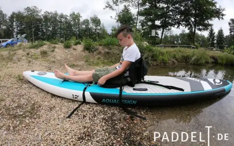 Junge sitzt auf SUP-Board, um den SUP Sitz zu befestigen. Das Board liegt am Ufer.
