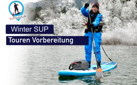 Person im blauen Neoprenanzug bereitet sich auf eine Winter SUP Tour vor. Die Landschaft ist mit Schnee bedeckt.