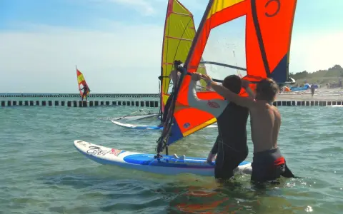 Zwei junge Männer bereiten sich auf das Windsurfen an der Ostsee vor. Ein weiterer Surfer ist in der Ferne zu sehen.