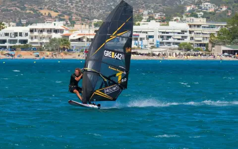 Windsurfer mit schwarzem Segel und gelben Akzenten auf türkisfarbenem Wasser vor einer Küste mit Gebäuden und Bergen. Surfen in Griechenland.