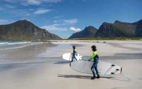 Zwei Surferinnen mit Neoprenanzügen und Surfbrettern am Strand in Norwegen. Die Berge im Hintergrund sind grün und die Wellen sind sanft.