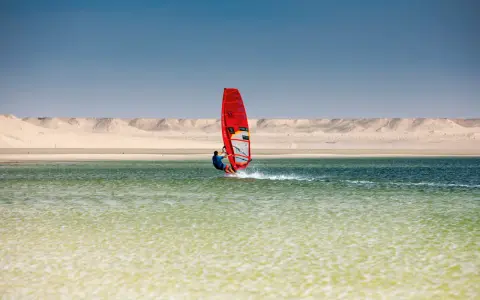 Windsurfer mit rotem Segel gleitet über das Wasser bei Dakhla, Marokko, vor einer Wüstenlandschaft.