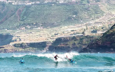 Surfer auf La Gomera reiten eine Welle, während im Hintergrund terrassierte Hänge und Häuser zu sehen sind.