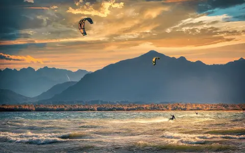 Kitesurfer gleiten über die Wellen am Chiemsee, während die Sonne hinter den Bergen untergeht. Ein traumhafter Abend für Windsurfen.
