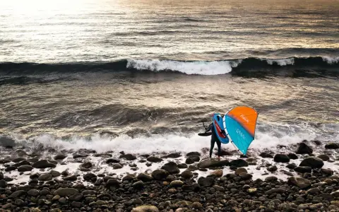 Ein Wingfoiler steht am steinigen Strand, bereit für den **shore break**. Die Sonne wirft ein warmes Licht auf die Wellen.