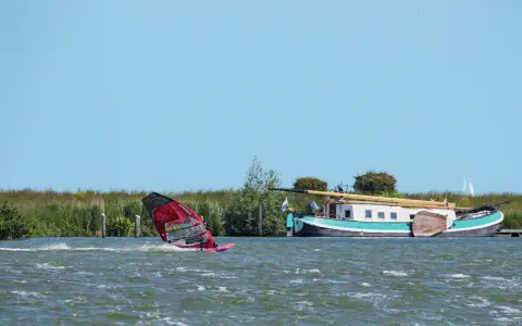 Windsurfen Holland: Ein Surfer gleitet über das Wasser, während ein Hausboot am Ufer liegt.