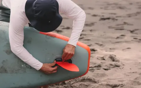 Mann mit Hut befestigt die rote Finne an einem aufblasbaren SUP-Board. Gute SUP-Pflege beginnt mit dem richtigen Zubehör.