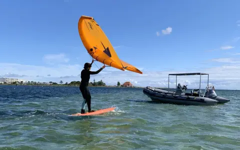 Person auf einem Board mit einem orangen Flügel, der über dem Wasser schwebt. Ein Schlauchboot liegt daneben. Foil surfen ohne Segel.