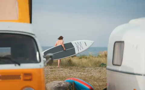 Mann mit SUP-Board am Strand von Fehmarn, bereit für die Wellen. Wohnmobile im Vordergrund.