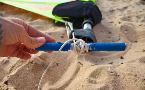 Hand hält blauen Griff, um das Segel zu spannen. Windsurf-Ausrüstung liegt im Sand.