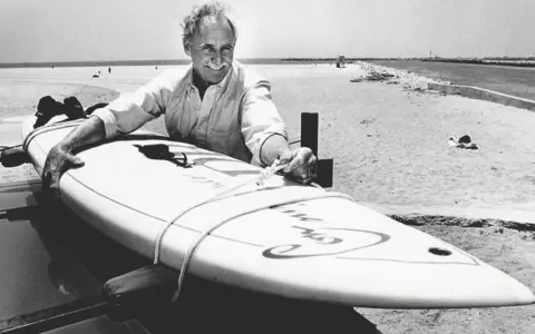 Jim Drake, ein Pionier des Surfens, lächelt stolz neben seinem Surfbrett am Strand.