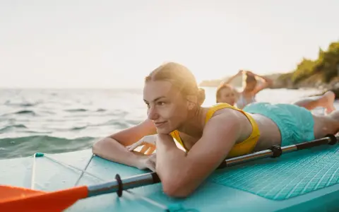 Junge Frau entspannt auf SUP-Board im Wasser. Ob Stand up paddling schwer ist, fragt sie sich lächelnd, während Freunde im Hintergrund planschen.