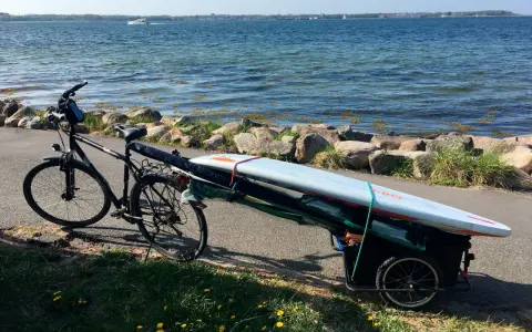 Fahrradanhänger mit Surfbrett und Paddel am Seeufer, bereit für ein Abenteuer auf dem Wasser.