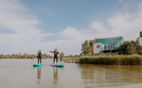 Zwei Frauen auf SUP-Boards am Makkum Strand, mit einem modernen Haus im Hintergrund.