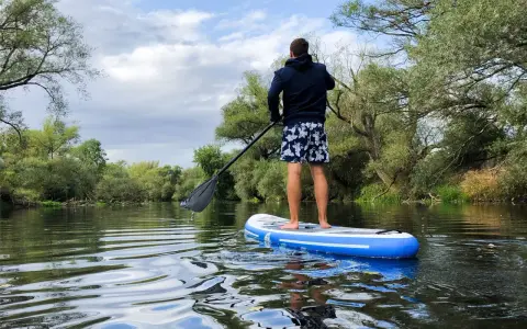 Mann auf SUP-Board auf ruhigem Fluss, umgeben von grünen Bäumen. Perfekt, um zu sehen, wo man stand up paddeln kann.