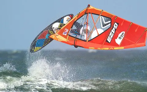 Lina Erpenstein fliegt mit ihrem Windsurf-Equipment über die Wellen. Das orangefarbene Segel und das bunte Board sind in der Luft.