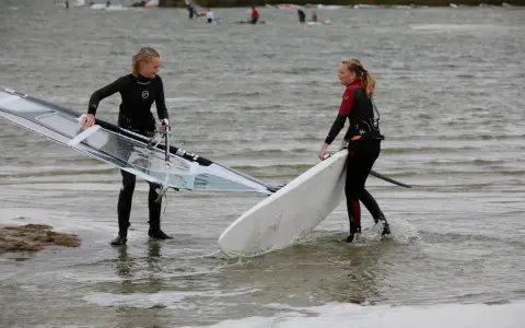 Zwei Mädchen bereiten sich auf das Windsurfen am IJsselmeer vor, tragen Neoprenanzüge und halten ihre Ausrüstung.